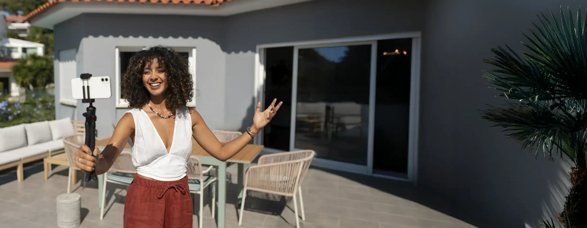 A woman with curly hair holds a black selfie stick with a white smartphone mounted on top.